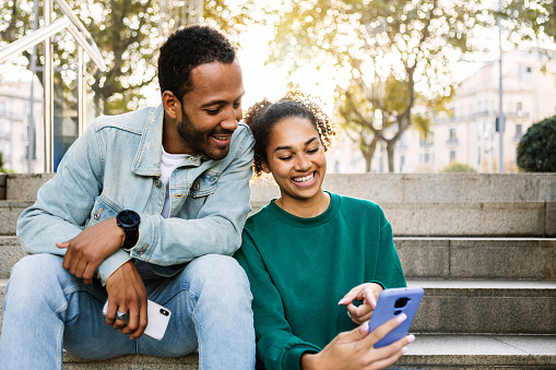 Two young latin american people using smart phone together sitting outdoors. Cheerful african american friends laughing while looking at cellphone screen. Social media concept. Copy space.