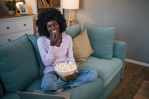 Beautiful young woman with an afro hairstyle watching movie in the night sitting on a couch in the living room at home and eating popcorn.