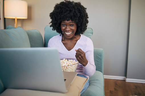 Beautiful young woman with an afro hairstyle watching movie. Sitting on a couch in the living room at home and eating popcorn.