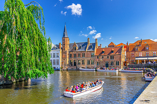 Bruges, Belgium, July 5, 2023: tourists boat in Dijver water canal of Reie river near Rosary Quay Rozenhoedkaai embankment, De groote Hollander Huidevettershuis in Brugge old town city centre