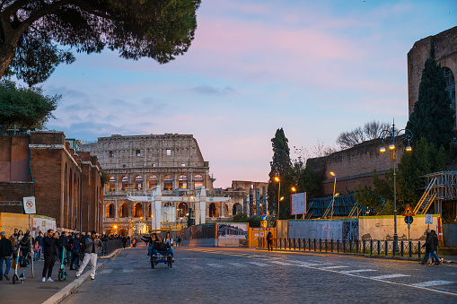 Rome, Italy - December 22, 2022: A bustling winter night in Rome, Italy, as people clad in mixed-use clothing walk and stand beneath the majestic Colosseum, surrounded by city lights and towering trees in the town square