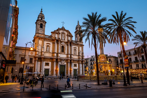 Palermo, Italy - December 24, 2022: A vibrant and bustling town square in san Domenico, Palermo, Italy, adorned with towering palm trees and a magnificent cathedral, all basking in the warm glow of a stunning blue hour. Nestled within the ancient quarter of La Loggia, this architectural masterpiece is a testament to Sicily's rich cultural heritage.