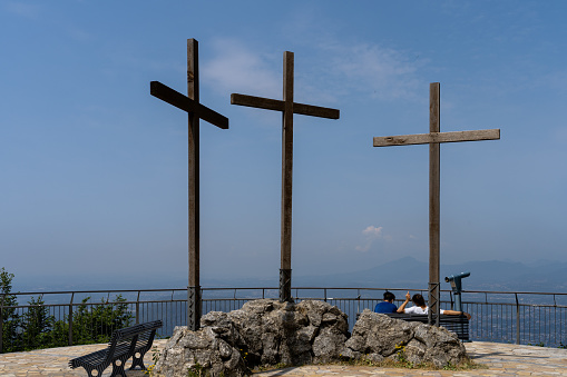 Brunate, Lombardy, Italy- June 27, 2023; With a magnificent view over Lake Como in Italy, and next to the Voltiano lighthouse a lookout point with three large bare wooden crosses, two boys rest sitting on a bench watching the scenery. Lighthouse on top of a hill, dedicated to the pioneer of electricity Alessandro Volta, overlooking the lake and the Alps.