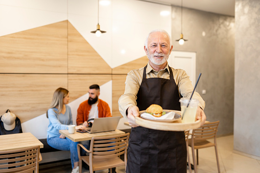Portrait of happy aged baker in apron standing against stall with bread in store