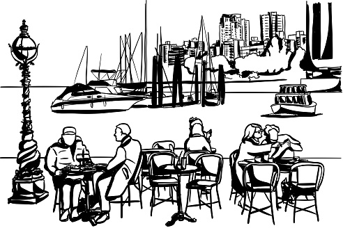 People sitting on a restaurant terrace by the waterfront.  Boats and city centre in the background.