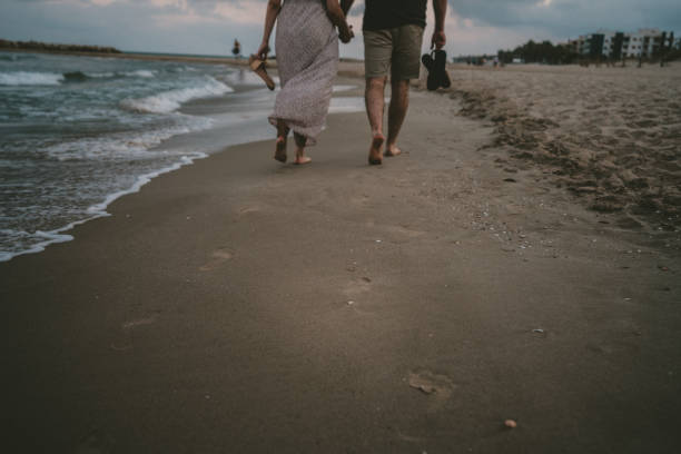 Couple walking along the shore of the beach with their backs to the camera. Footprint marks in the sand on the shore with gentle waves of the sea next to it. They hold their flip flops in their hands.