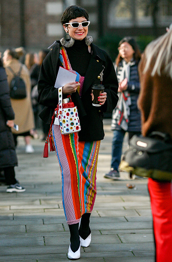 London, UK 02 17 2019 Photo of woman with short hair and sunglasses  wearing a  vibrant colourful outfit including multi coloured  pattern striped skirt, black coat, white stilettos heels shoes walking outdoors on the street