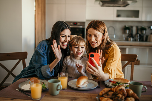 Same-sex couple and their adopted daughter share a meal and laughter while using a phone for a video call