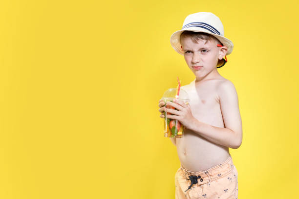 Cute boy in panama drinking Fresh juice from plastic cup over yellow studio background