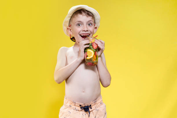 Cute boy in panama drinking Fresh juice from plastic cup over yellow studio background