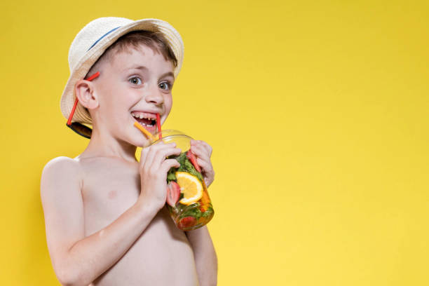 Cute boy in panama drinking Fresh juice from plastic cup over yellow studio background