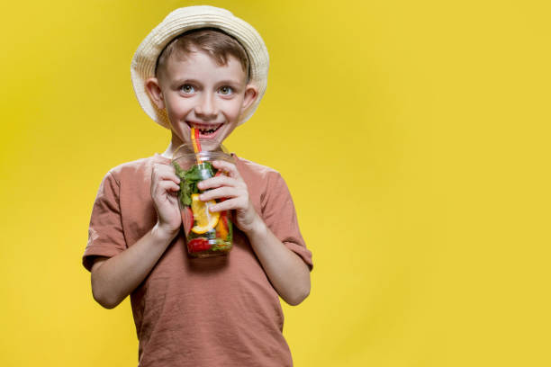 Cute boy in panama drinking Fresh juice from plastic cup over yellow studio background
