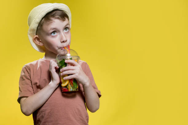 Cute boy in panama drinking Fresh juice from plastic cup over yellow studio background