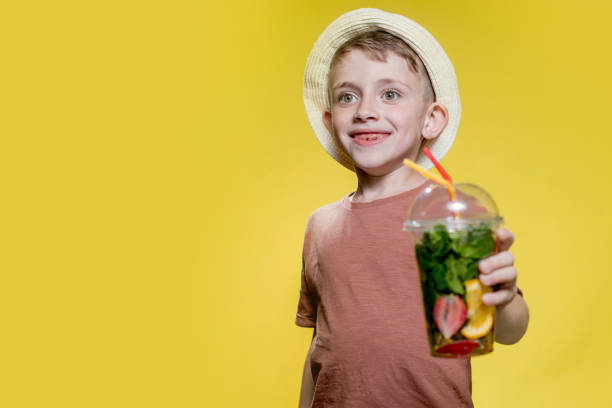 Cute boy in panama drinking Fresh juice from plastic cup over yellow studio background