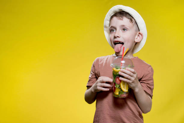 Cute boy in panama drinking Fresh juice from plastic cup over yellow studio background