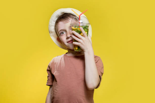 Cute boy in panama drinking Fresh juice from plastic cup over yellow studio background