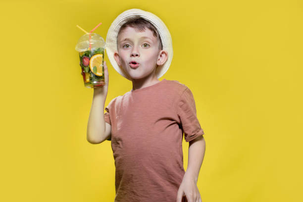 Cute boy in panama drinking Fresh juice from plastic cup over yellow studio background