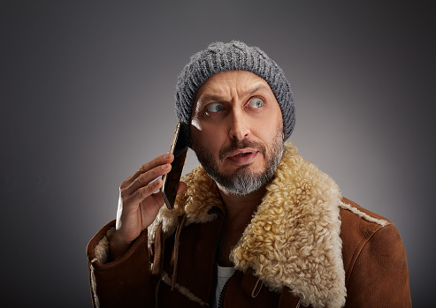 Man wearing leather coat with fur collar and knit hat. Man with big eyes and curious expression talking on the phone