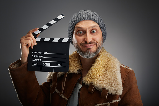 Man wearing leather coat with fur collar and knit hat looking with big smile and big eyes with a clapperboard