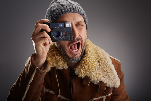 Man wearing leather coat with fur collar and knit hat super is excited while taking a picture with his old digital camera