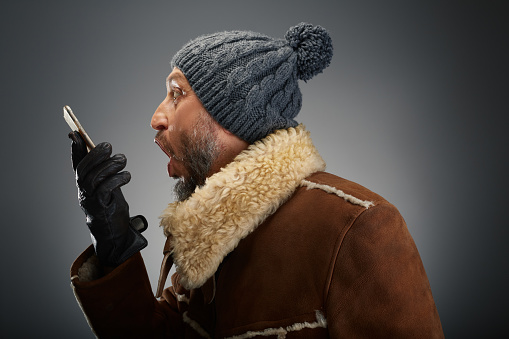 Man wearing leather coat with fur collar and knit hat making funny expressions while talking on the phone