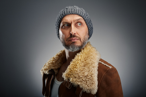 Handsome man wearing leather coat with fur collar and knit hat looking away with raised eyebrow