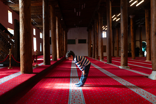The boy praying in the mosque bows. Wooden poles serve as columns inside the mosque. Mosque built of wood and stone. Prayer is performed on the red carpet. Taken in daylight with a full frame camera.