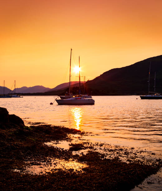 An image capturing the serene beauty of Glencoe near the sea at sunset, with the warm hues of the evening sky reflecting on the water. In the foreground, moored boats and yachts rest peacefully, set against the dramatic backdrop of the Scottish Highlands, creating a picturesque and tranquil maritime scene.