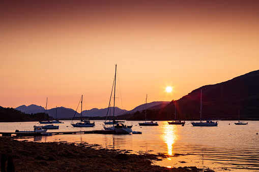 An image capturing the serene beauty of Glencoe near the sea at sunset, with the warm hues of the evening sky reflecting on the water. In the foreground, moored boats and yachts rest peacefully, set against the dramatic backdrop of the Scottish Highlands, creating a picturesque and tranquil maritime scene.