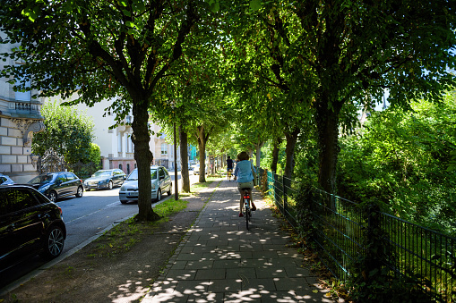 Strasbourg, France - July 29, 2017: Rear view of single woman cycling on the French trottoir under big canopies of green trees