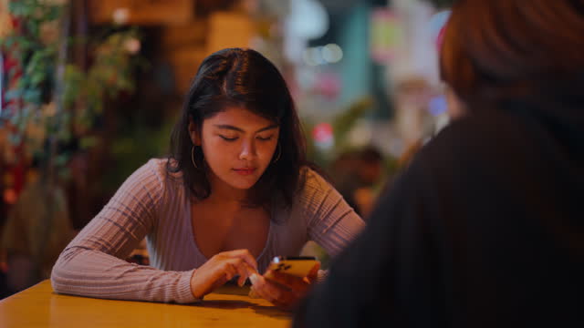 Two multiethnic female tourist friends using their mobile smart phones and enjoying talking with each other in sidewalk cafe at night