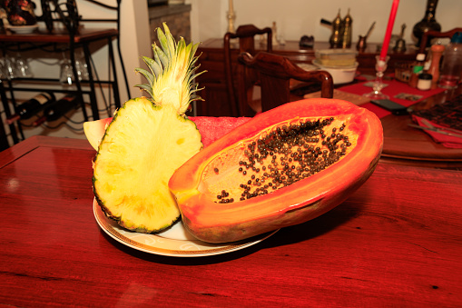 Half a ripe pineapple and papaya on a plate on a shiny rosewood table. In the background, there is a segment of watermelon. They are ready to eat.