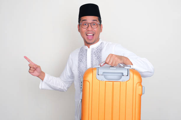 istock Moslem Asian man carry a suitcase and pointing to the right side with excited expression 2006290486
