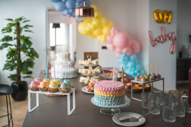 A baby shower table adorned with a multi-tiered rainbow cake, elegant snacks, and a vintage-style drink dispenser in a contemporary space.