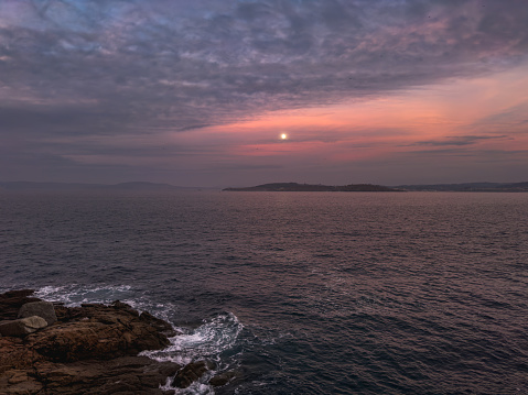 the ethereal beauty of a moonrise over a calm sea, as twilight paints the sky in shades of purple and orange. The rugged coastline, marked by craggy rocks and frothy waves, contrasts with the smooth, dusky horizon where the sea meets the sky. The distant moon, a glowing orb suspended in the gradient twilight, casts a subtle light over the landscape, creating a serene and contemplative atmosphere. This moment, where day gently succumbs to night, is a testament to nature's quiet grandeur.