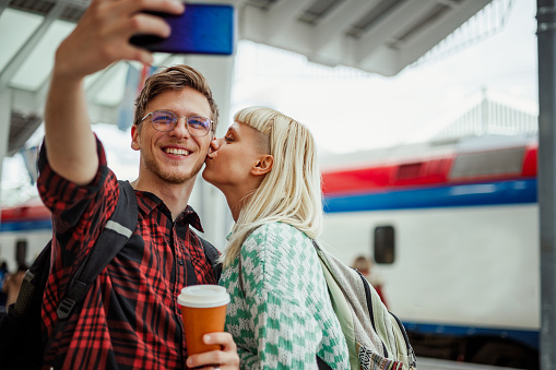 Young happy couple at the train station is waiting for their train to arrive while taking a selfie. Woman is kissing a man while he is holding a coffee takeaway and taking photo of the two of them.