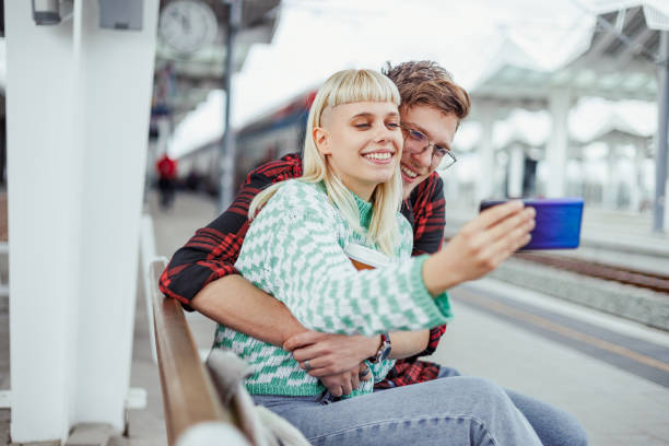 istock Young couple taking a selfie while hugging and sitting on a bench at the railway 2006327928