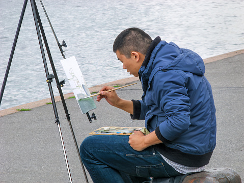 St. Petersburg, Russia - July 2, 2014: Young artist with portable easel in warm jacket and jeans paints  landscape on Neva River embankment near Church of Assumption of Mary on Vasilyevsky Island, Lieutenant Schmidt Embankment, Saint Petersburg, Russia