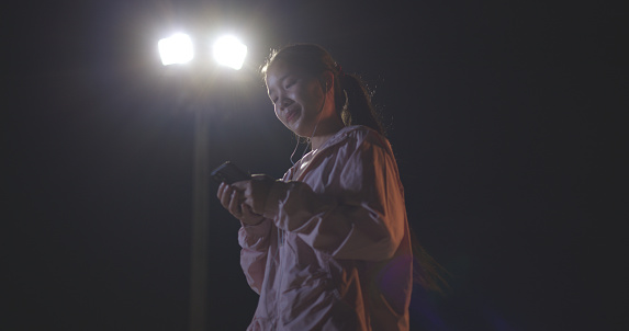 Selective focus, low angle view, Young female runner in sportswear is having break and enjoy listening to music with headphones on mobile phone at night time.preparing for evening workout.healthy lifestyle.