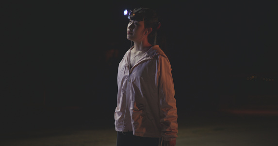 Side view, Young female runner in sportswear use flashlight on head and listening to music with headphones while exercising in the outdoor sports stadium at night time.preparing for evening workout.healthy lifestyle.