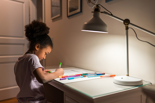 A concentrated mixed-race girl engages in homework activities at a well-lit home desk, with her Caucasian father assisting. Both are in a comfortable indoor setting, emphasizing a supportive educational environment.