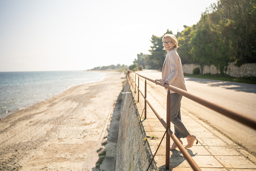 Full length shot of mature businesswoman enjoying sea view while leaning on promenade fence in coastal town