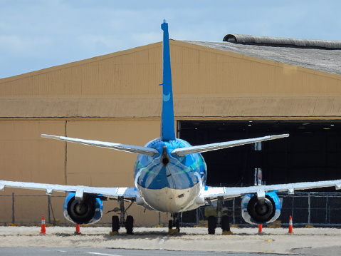 A Nauru Airlines Boeing B737-319(SF) plane, registration VH-VNU, parked behind the domestic terminal of Sydney Kingsford-Smith Airport. In the background is a hangar. This image was taken from Ross Smith Avenue, Mascot on a hot, sunny and partly cloudy afternoon on 10 February 2024.