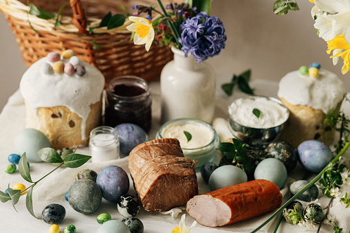 Happy Easter! Stylish easter natural dyed eggs, meat, bread, butter, beets, basket and flowers on rustic table. Traditional easter orthodox holiday food for blessing and daffodils bouquet