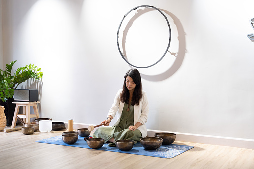 A young woman of Asian descent sits on a wooden floor, engaging with a singing bowl instrument