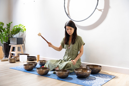 An Asian young woman sits on a wooden floor, her focus solely on the singing bowl instrument in her hands