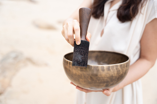 Presented in a portrait a young Asian woman holds a singing bowl instrument while standing on a white sand beach