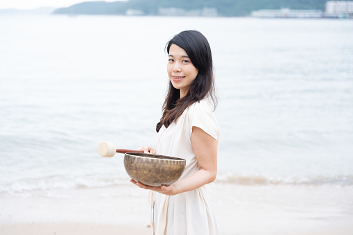 A young Asian woman portrayed in a portrait holding a singing bowl instrument on a white sand beach