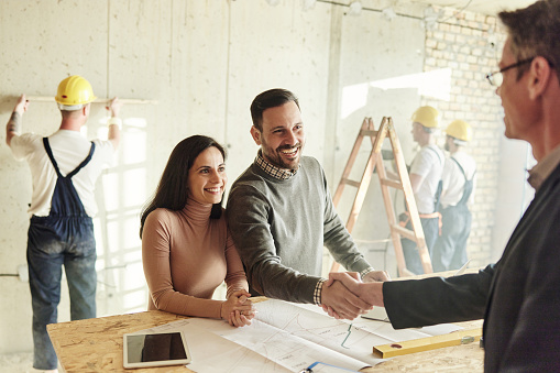 Happy couple and home designer came to a successful agreement at construction site. Manual workers are in the background.