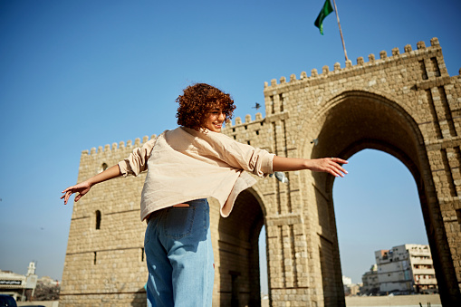 Smiling woman twirling with arms outstretched in front of famous arched gateway marking the start of pilgrimage route to Mecca.